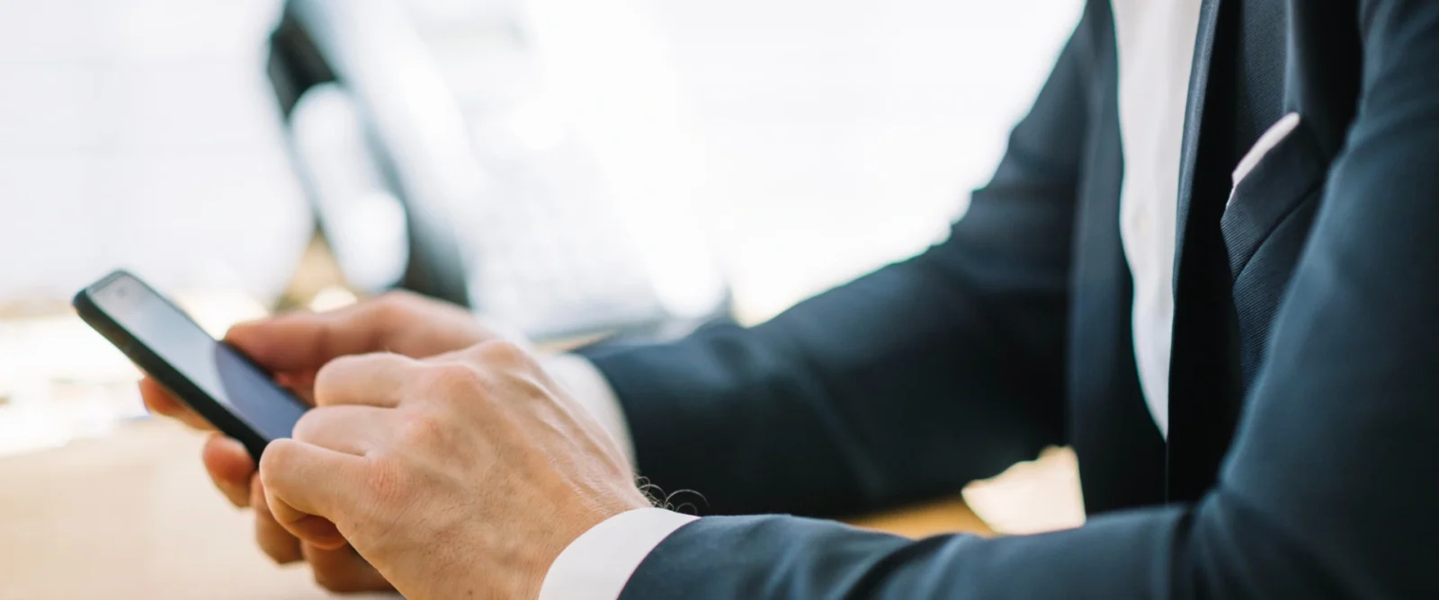 An eye-level close-up shot of a person dressed in a dark suit with a white shirt, holding a smartphone in both hands and typing on it. The background is a bright, blurred office environment.